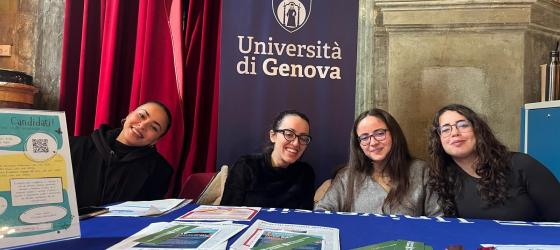 tavolo informativo con depliant e 4 giovani donne sullo fondo uno stendardo unige