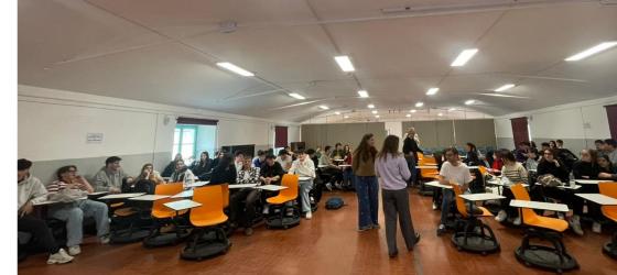vista aula scolastica con una serie di studenti e studnetesse sedute ai banchi e alcune persone che parlano al centro dell'aula