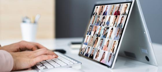 in primo piano una mano che scrive su un pc, sullo schermo si vedono motle persone collegate in videoconferenza