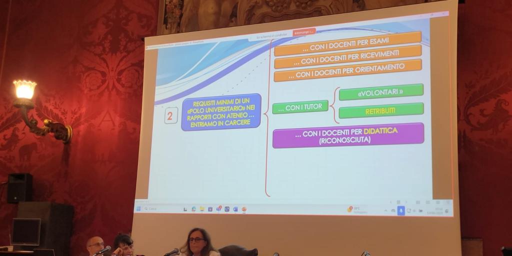 immagine interno sede conferenza con sfondo slide proiettata e relatore al tevolo